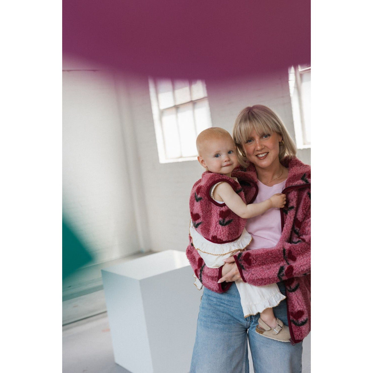 Lulu Baby bodywarmer cherry wordt gedragen door vrouw die baby vasthoudt in licht interieur met ramen