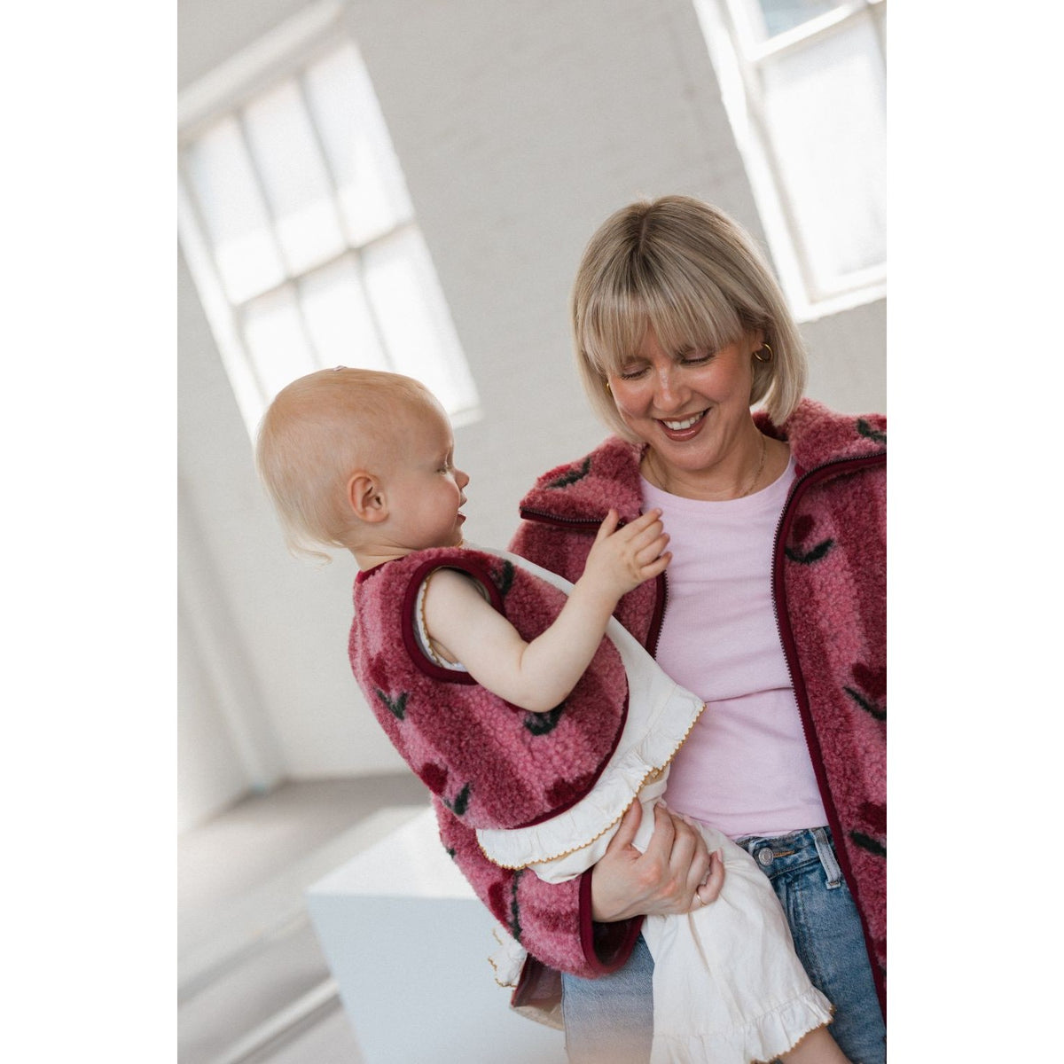 Lulu Baby bodywarmer in cherry rood, gedragen door peuter naast volwassene in helder interieur met ramen