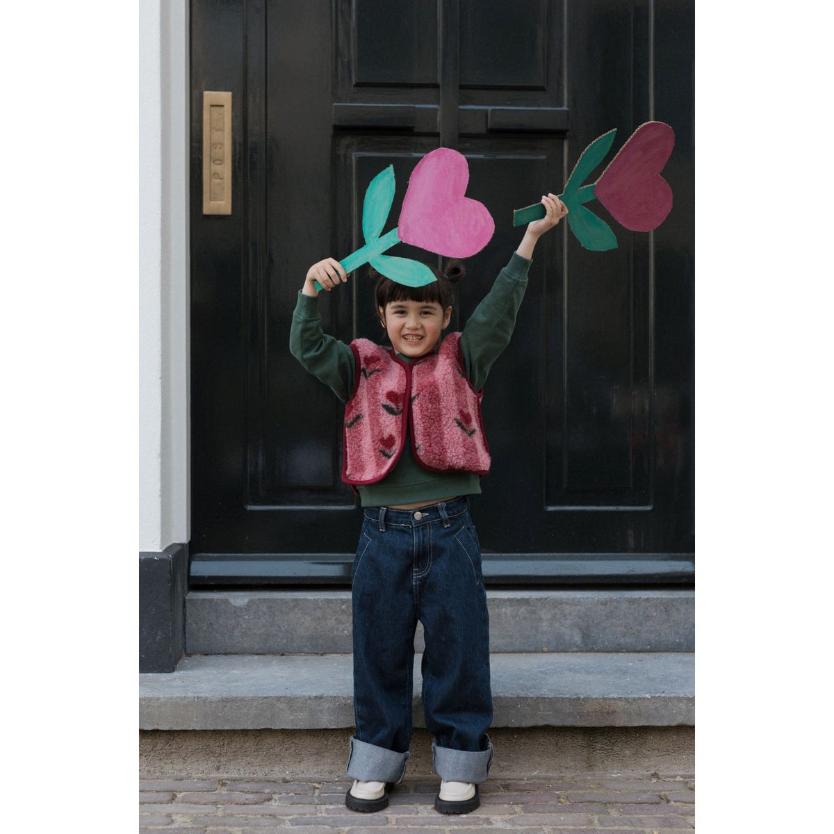 Alwero Camino gilet cherry: kind draagt roze quilted gilet over groen shirt, speelnd met gekleurde papieren bloemen voor zwarte deur.