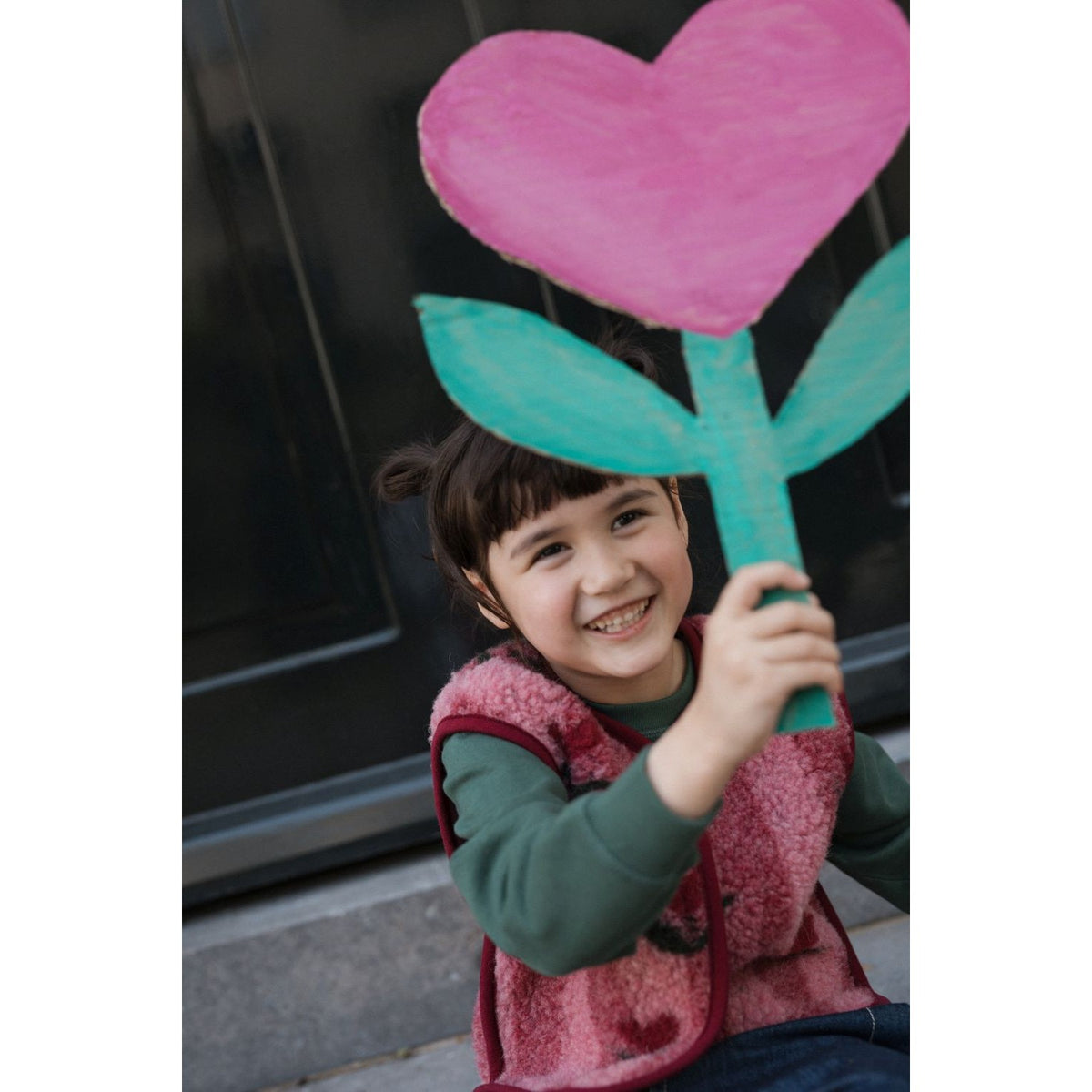 Alwero Gilet Camino junior Cherry: kind draagt roze bodywarmer met groen shirt, lachend met groot hart en turquoise stengel boven hoofd