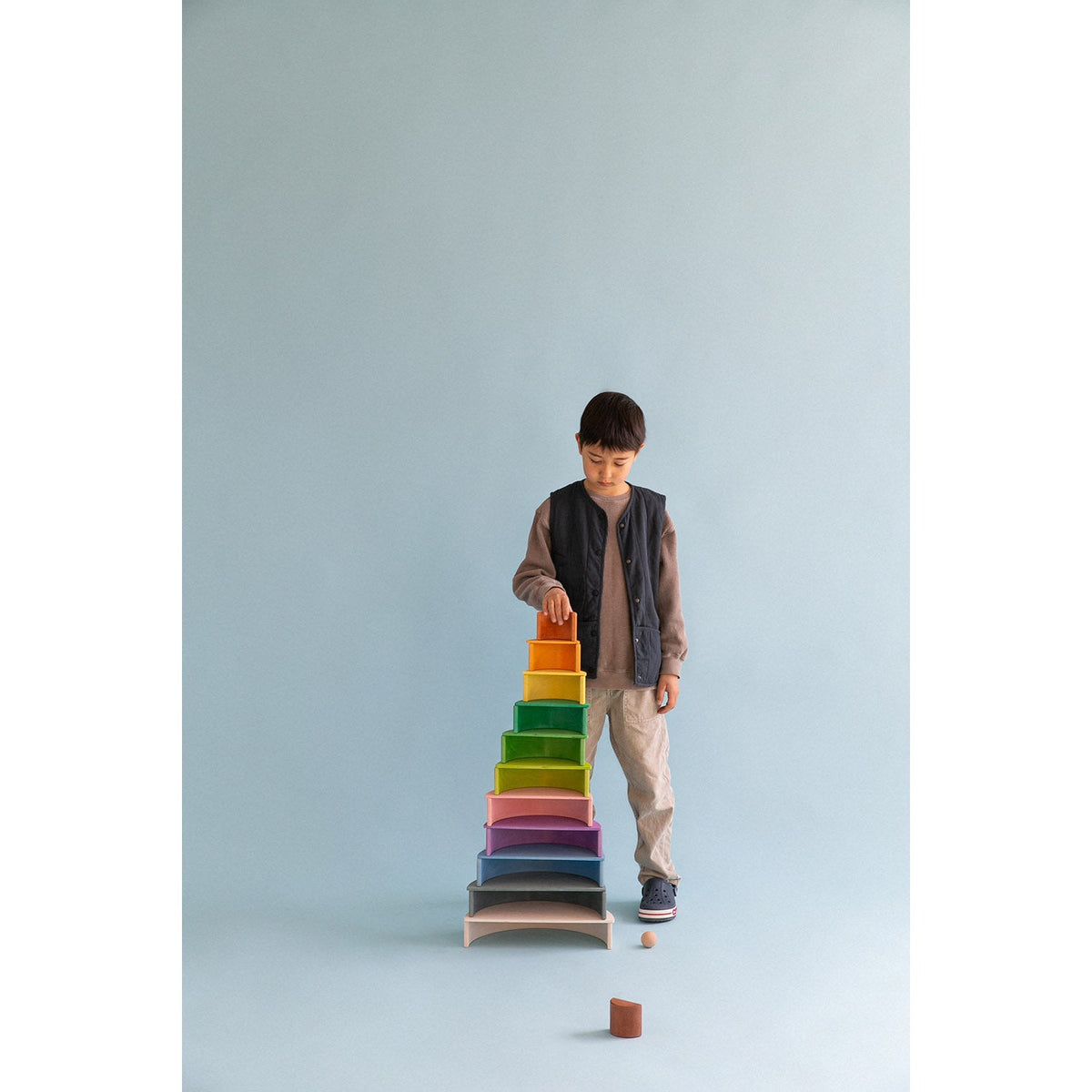 Avdar Rainbow Stacker Large: jongen bouwt stapeltoren met gekleurde houten bogen tegen blauwe achtergrond