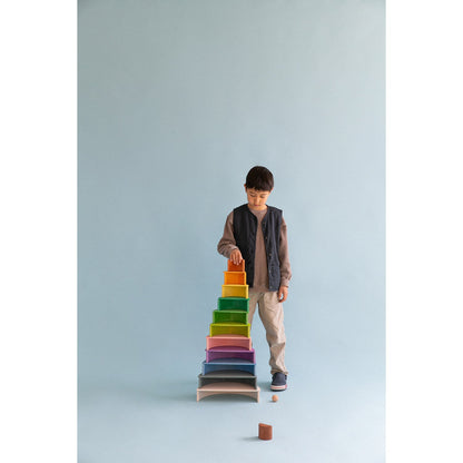 Avdar Rainbow Stacker Large: jongen bouwt stapeltoren met gekleurde houten bogen tegen blauwe achtergrond