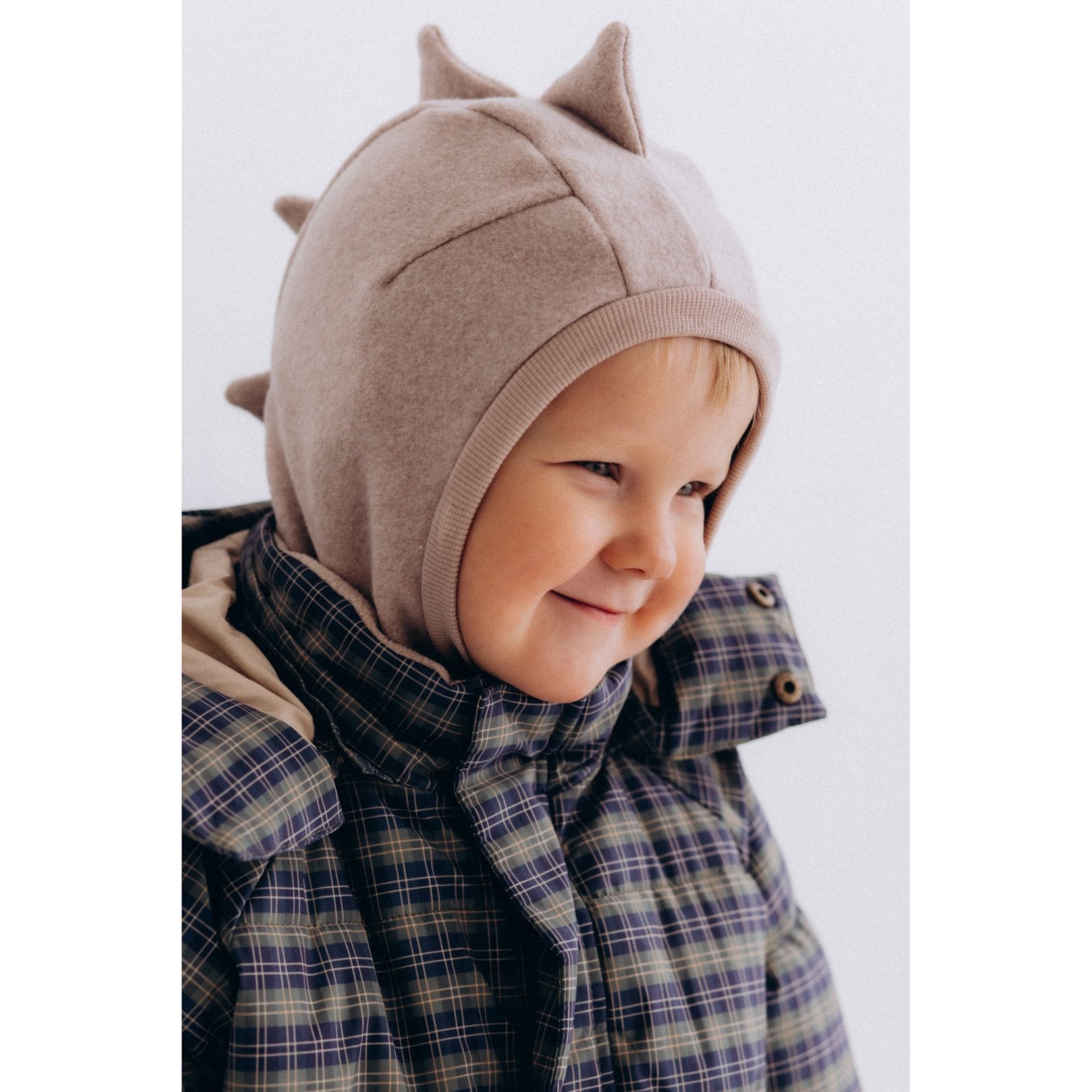 Wooly Organic balaclava dino in lichtgrijs met speelse dinosaurusspikes, uitgeprobeerd door peuter in geruit jasje