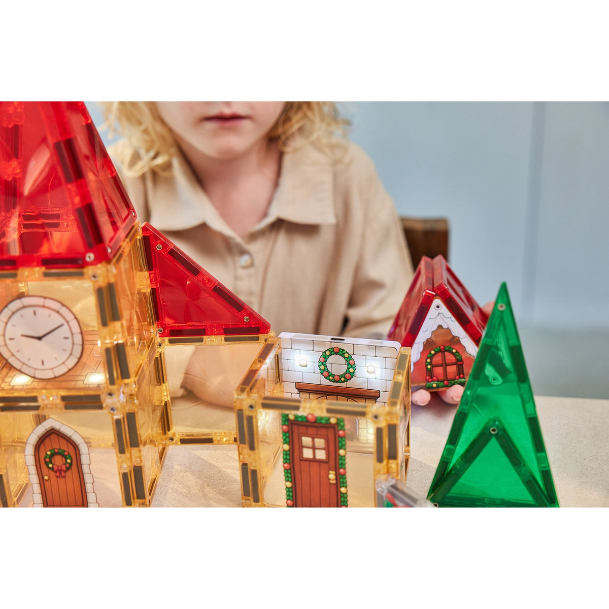 Cleverclixx Christmas Village adventskalender met magnetische tegels: kind bouwt verlichte kersthuizen met klokketoren, groene bomen en rode daken.