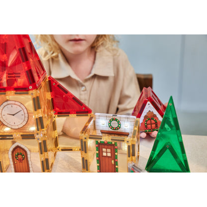 Cleverclixx Christmas Village adventskalender met magnetische tegels: kind bouwt verlichte kersthuizen met klokketoren, groene bomen en rode daken.
