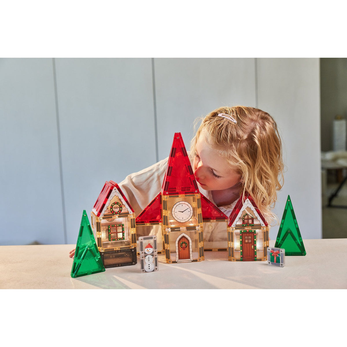 Cleverclixx kerstdorp adventskalender met magnetische tegels: meisje speelt met verlichte kerkgebouwen, groene bomen en sneeuwpop.