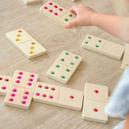 Tickit houten edelsteen domino: kinderhand plaatst gekleurde juweeltjes-dominosteen, meerdere stenen zichtbaar op houten tafel.