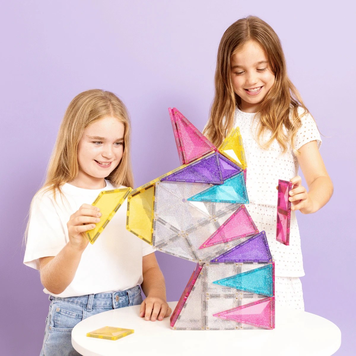 Connetix magnetische glitter tegels: twee meisjes bouwen een torenvormige structuur met gekleurde driehoekige stenen tegen paarse achtergrond.