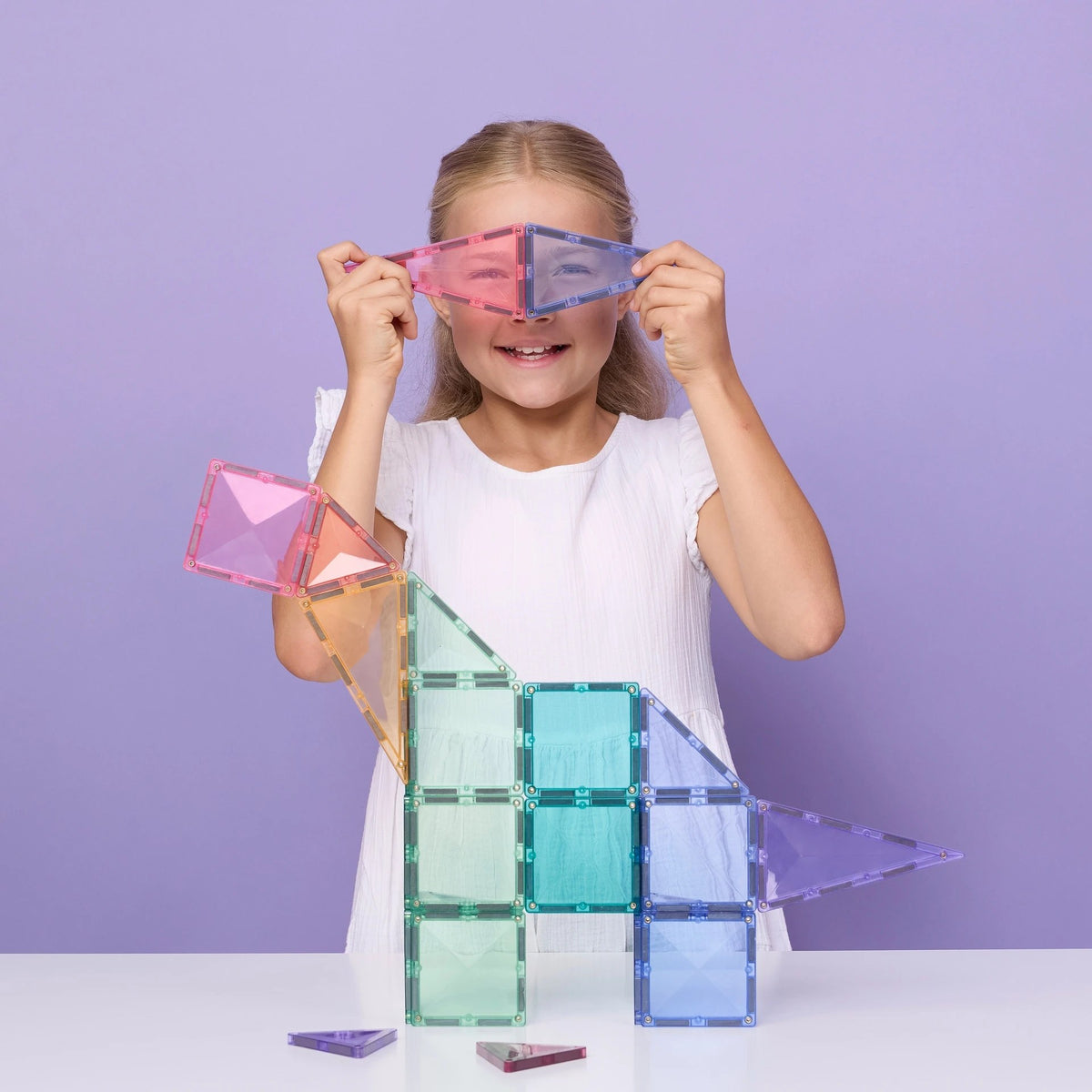 Meisje speelt met Connetix Tiles magnetische bouwstenen in pastelkleuren, bouwt een huis en houdt gekleurde bril omhoog