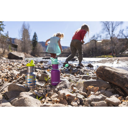 Klean Kanteen kinderfles 355ml met sippy cap in vooraanzicht op stenen strand aan rivier, kinderen spelend op achtergrond