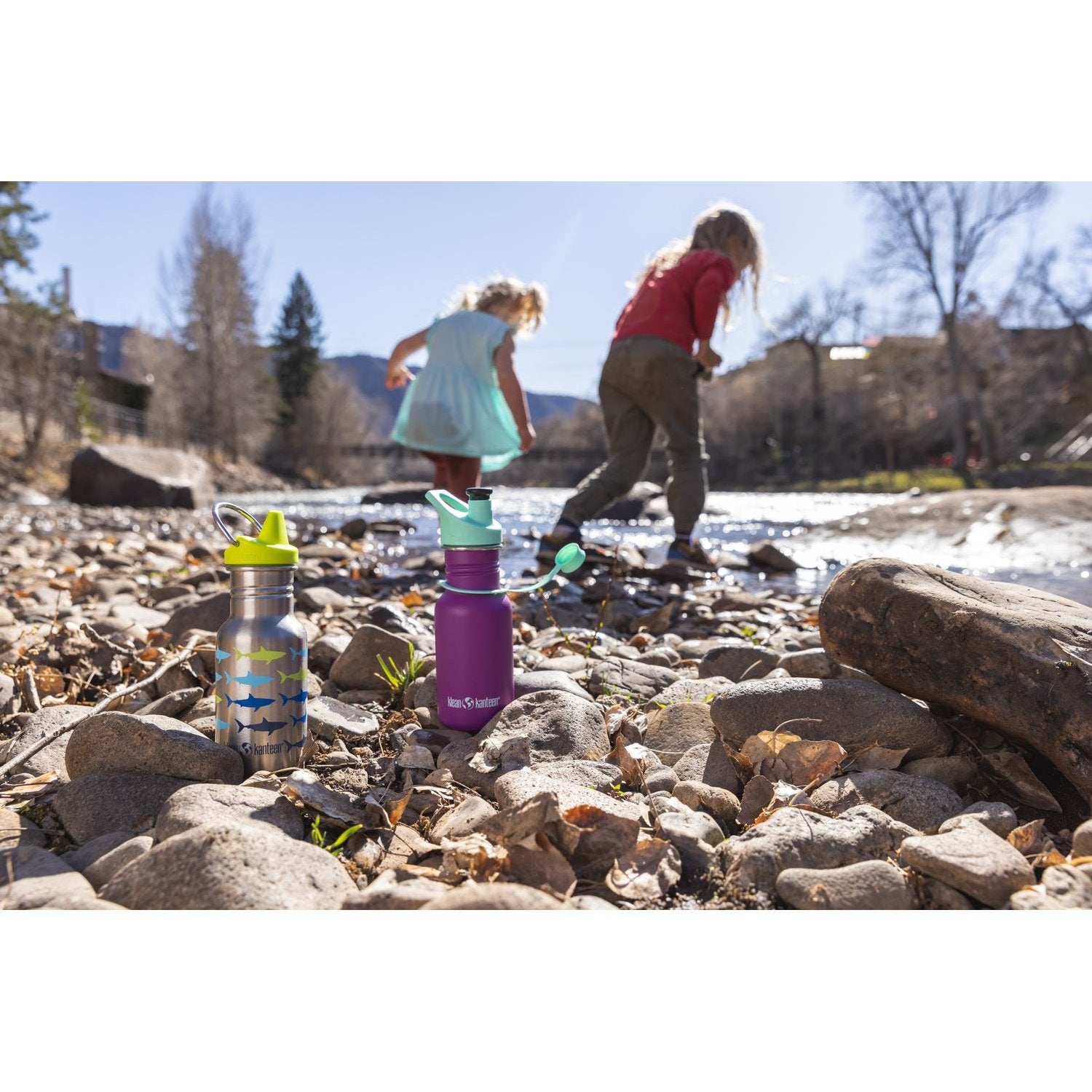 Klean Kanteen Classic 355ml drinkfles met sippy cup voorgrond op stenen strand, kinderen spelen achter bij rivier