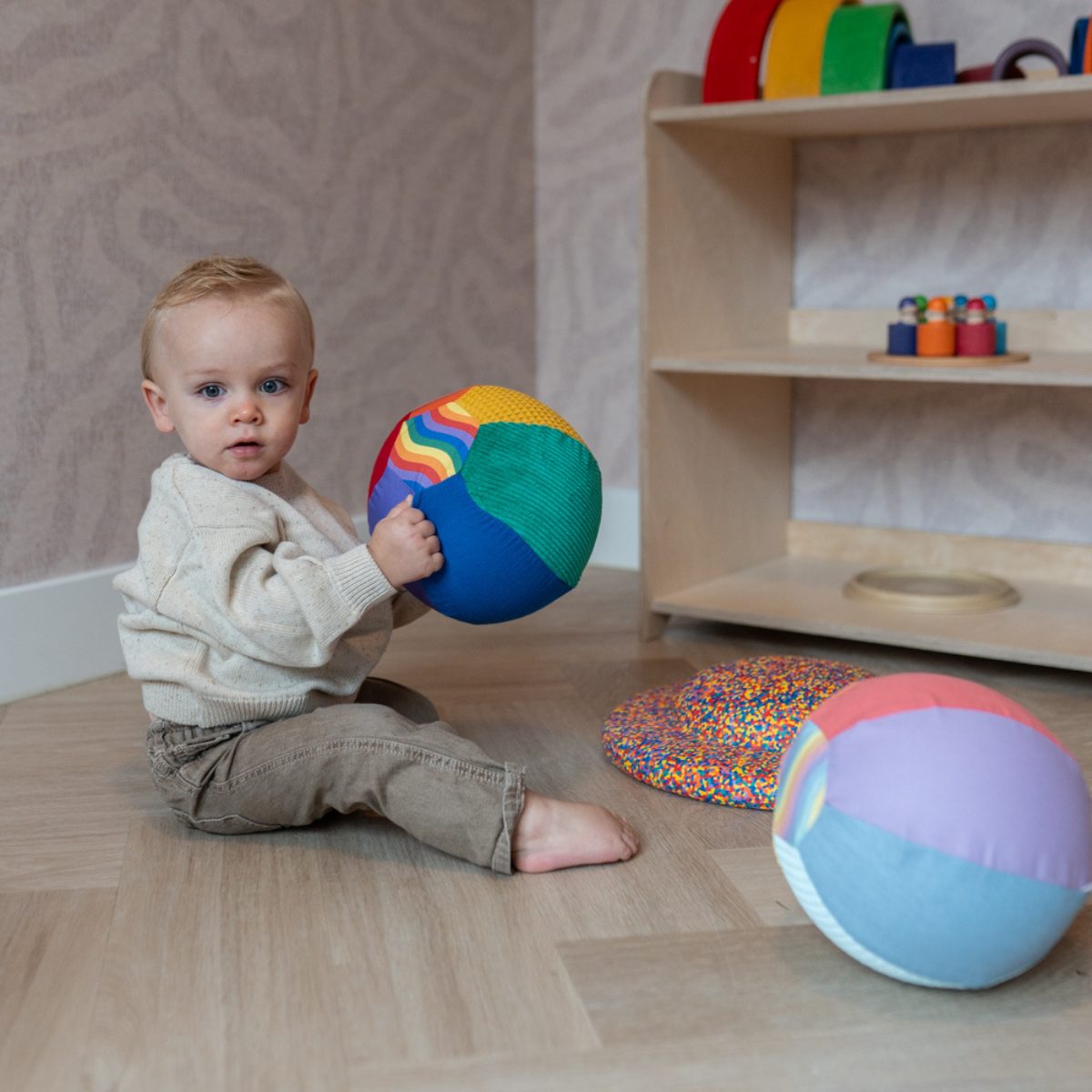 Little Botanic Label sensorische ballonbal in groen en blauw wordt vastgehouden door zittend peuter, met speelballen op houten vloer