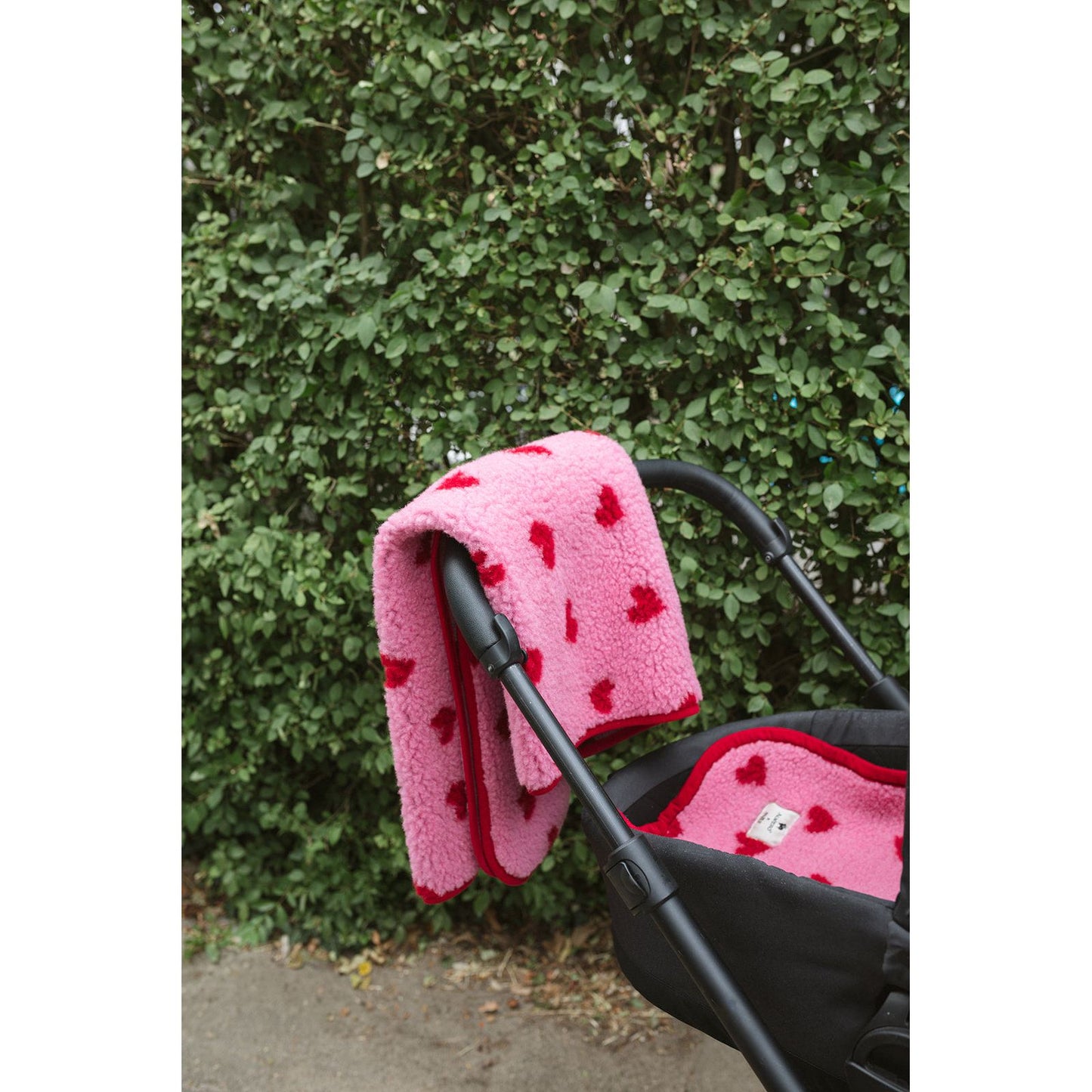 Alwero wiegbeschermer in roze wol met rode hartjes, draped over kinderwagen tegen groene haag