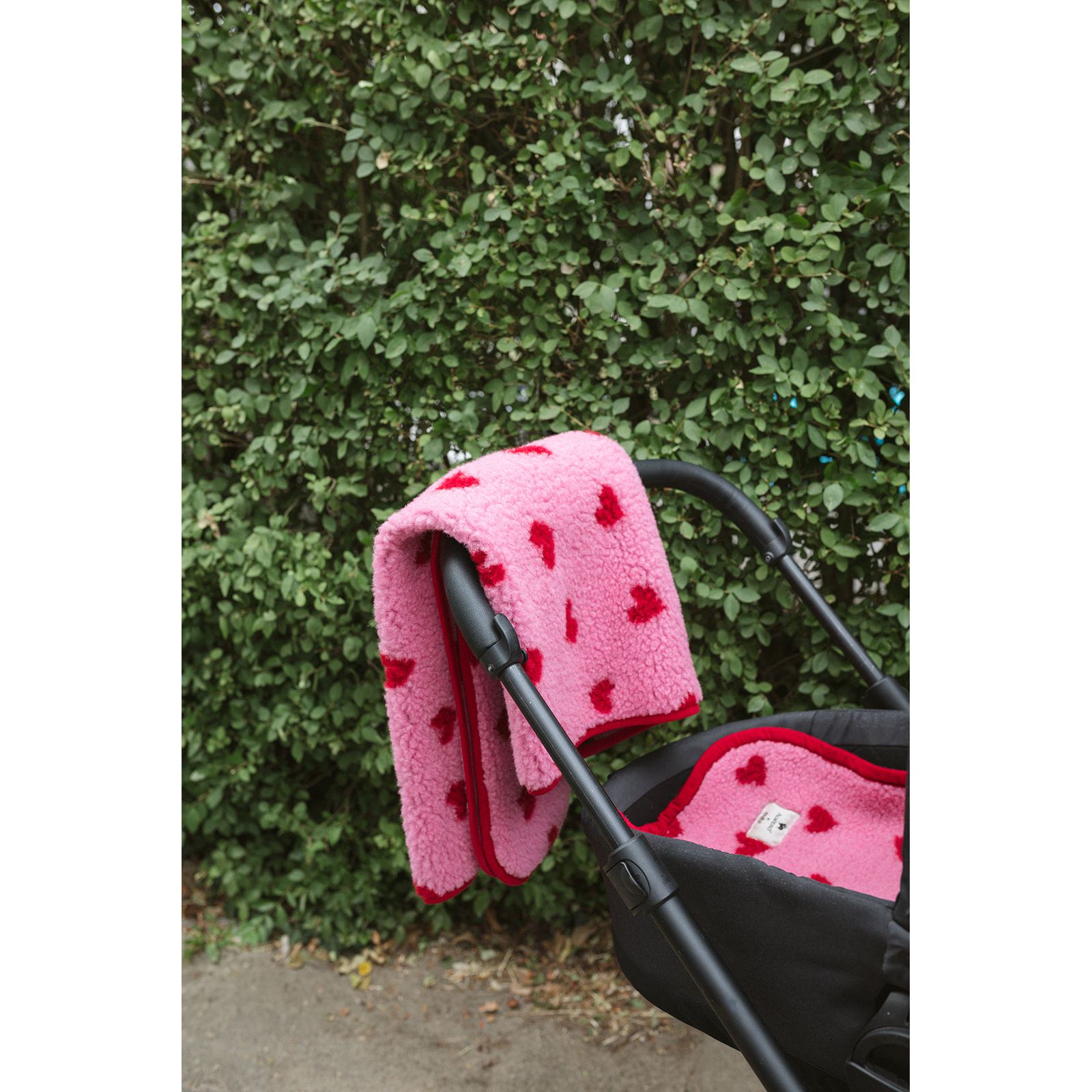 Alwero wiegbeschermer in roze wol met rode hartjes, draped over kinderwagen tegen groene haag