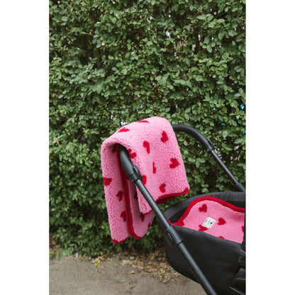 Alwero wiegbeschermer in roze wol met rode hartjes, draped over kinderwagen tegen groene haag
