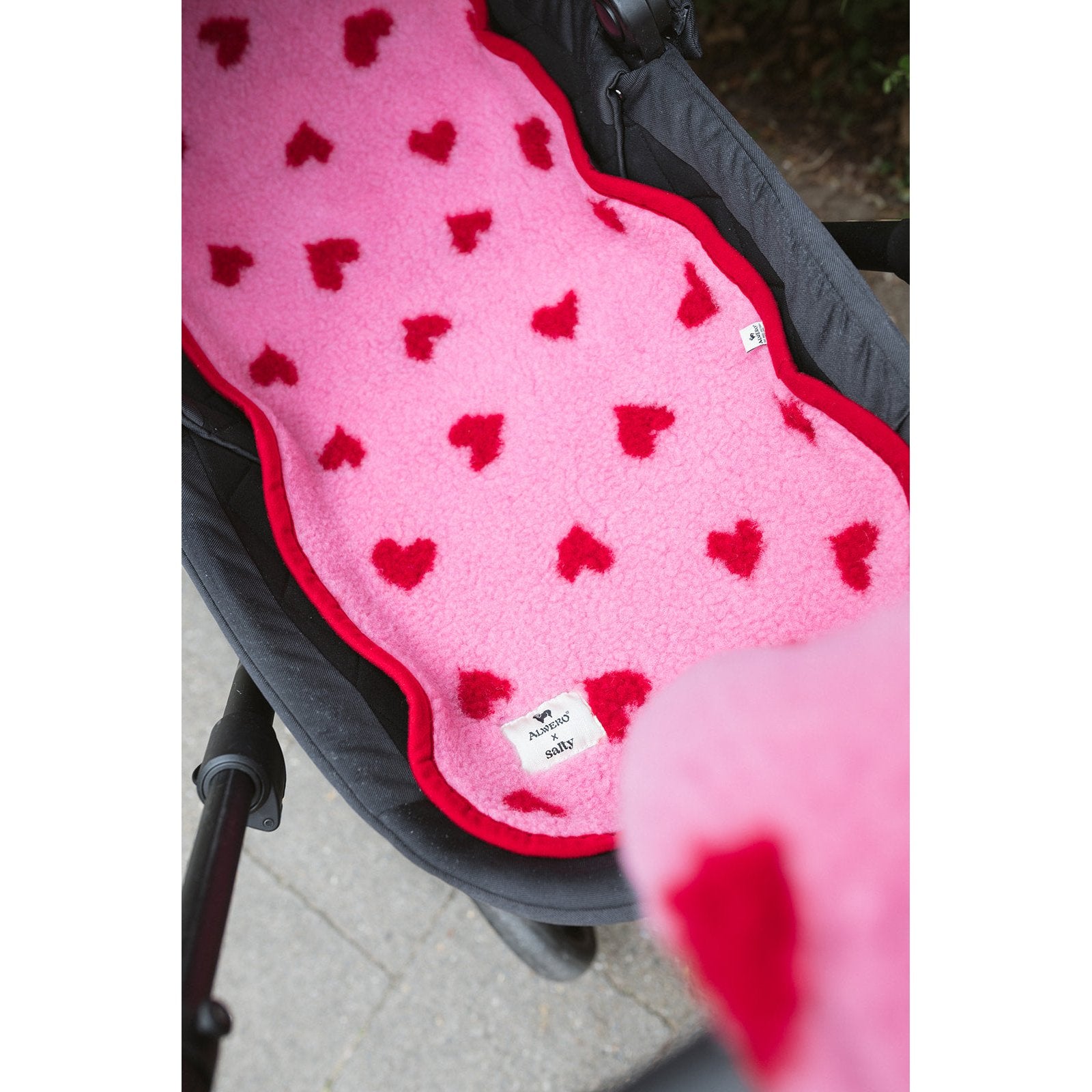 Alwero wiegbeschermer in roze wol met rode hartjes, uitgespreid over zwarte kinderwagen vanuit bovenaanzicht