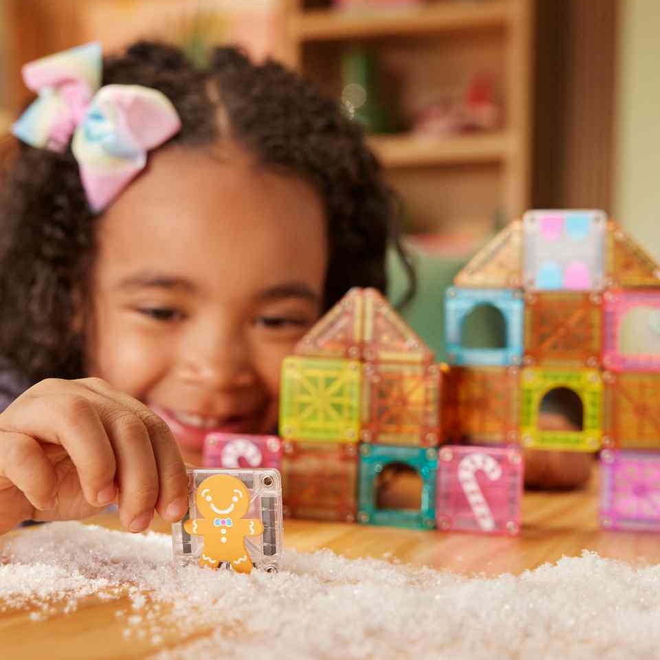 Magna-Tiles microMAGS Gingerbread House set: kind bouwt met kleurrijke magnetische tegels, gingerbread-figuur in voorgrond op zand