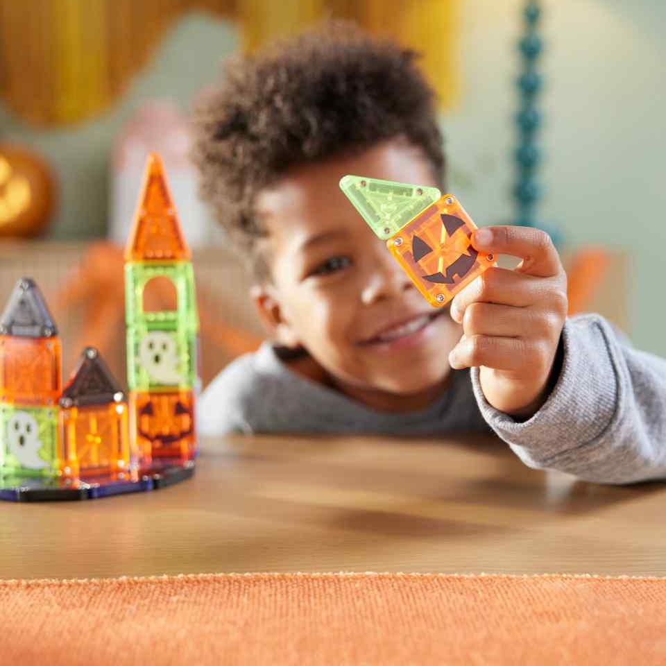 Kind speelt met Magna-Tiles Travel set magnetische bouwstenen, houdt oranje pompoen-element omhoog naast groen kasteel