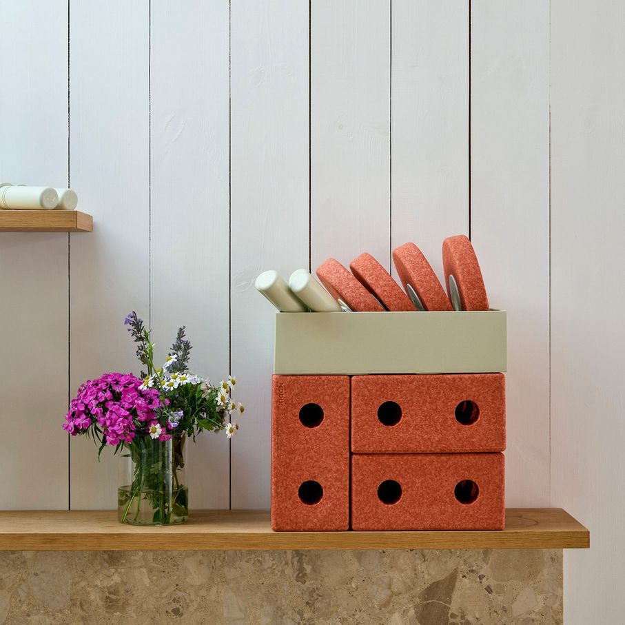 MODU 3-in-1 Baby Walker in Burnt Orange met modulaire blokken op houten tafel, gevuld met rollen en handdoeken, naast bloemen