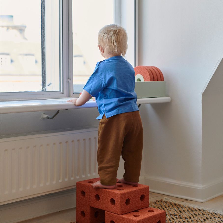 MODU 3-in-1 Baby Walker in Burnt Orange als opstapje bij raam, peuter staat op modulaire blokken om naar buiten te kijken