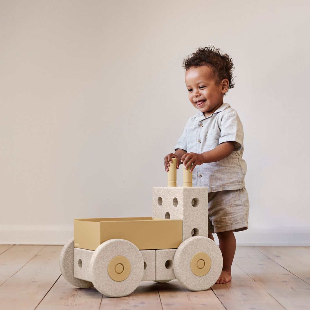 MODU 3-in-1 babywalker in sand grey en honey yellow met peuter die ermee loopt, vooraanzicht op houten vloer