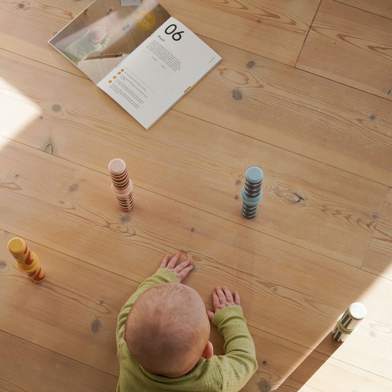 MODU Baby Activity Set Burnt Orange Dusty Green - bovenaanzicht van baby in groene trui die speelelementen onderzoekt op houten vloer met instructieka