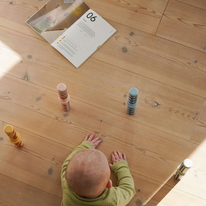 MODU Baby Activity Set Burnt Orange Dusty Green - bovenaanzicht van baby in groene trui die speelelementen onderzoekt op houten vloer met instructieka