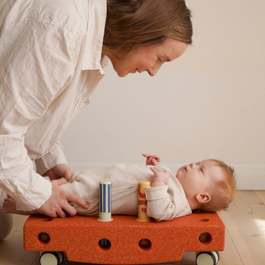 MODU Baby Activity Set burnt orange - moeder ondersteunt baby liggend op speelbord met rammelaars en textuurblokken