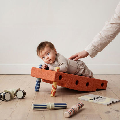 MODU Baby Activity Set Burnt Orange met baby die op balansbord ligt, hand van volwassene ondersteunt, speelgoed rond op houten vloer