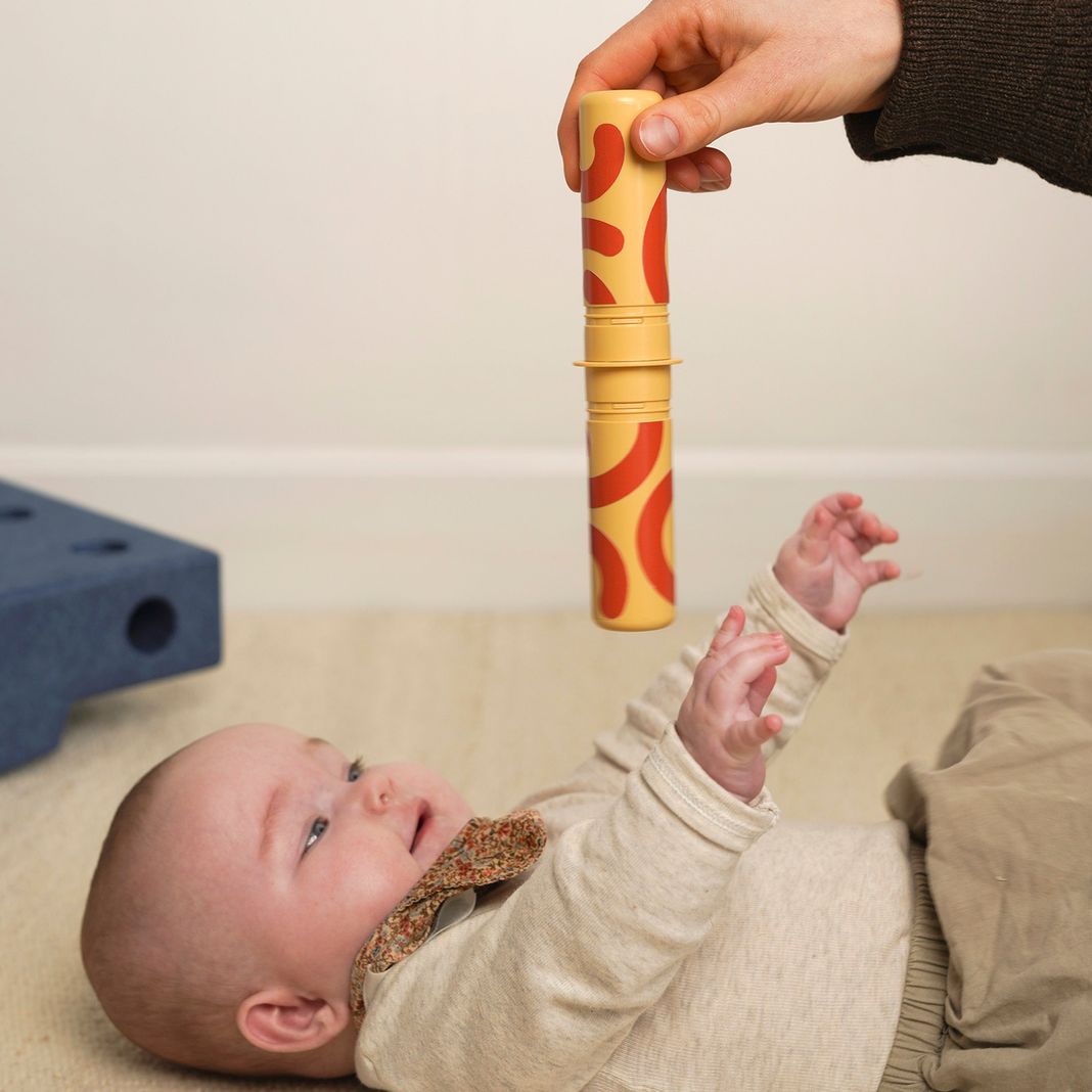 MODU Baby Activity Set: volwassene houdt geel speeltuig met rode details boven baby die er naar kijkt