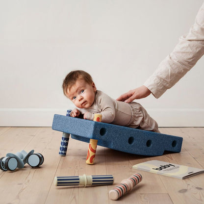 MODU Baby Activity Set Deep Blue met baby liggend op blauwe speelbalk, omringd door speelgoed onderdelen, hand van volwassene ondersteunend.