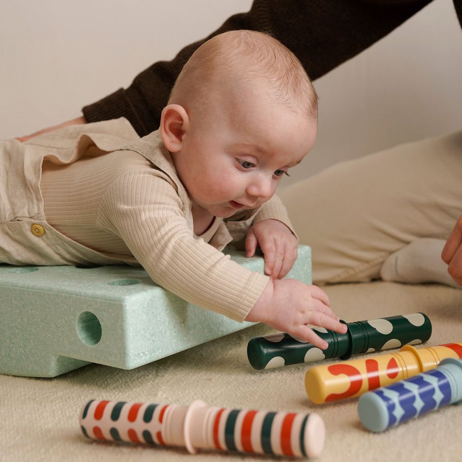 Baby speelt met gekleurde MODU speelrollen naast groen schuimblok, ondersteund door volwassene