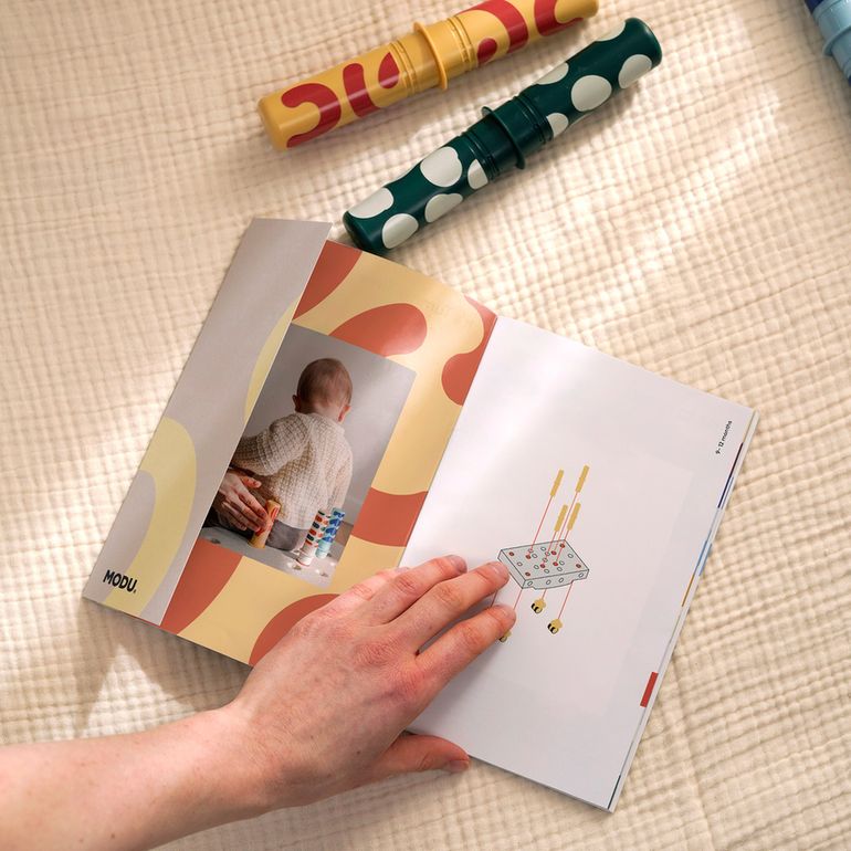 MODU Baby Activity Set met kleurrijke speelstaven en instructieboekje, hand houdt open gids vast op witte gewatteerde ondergrond