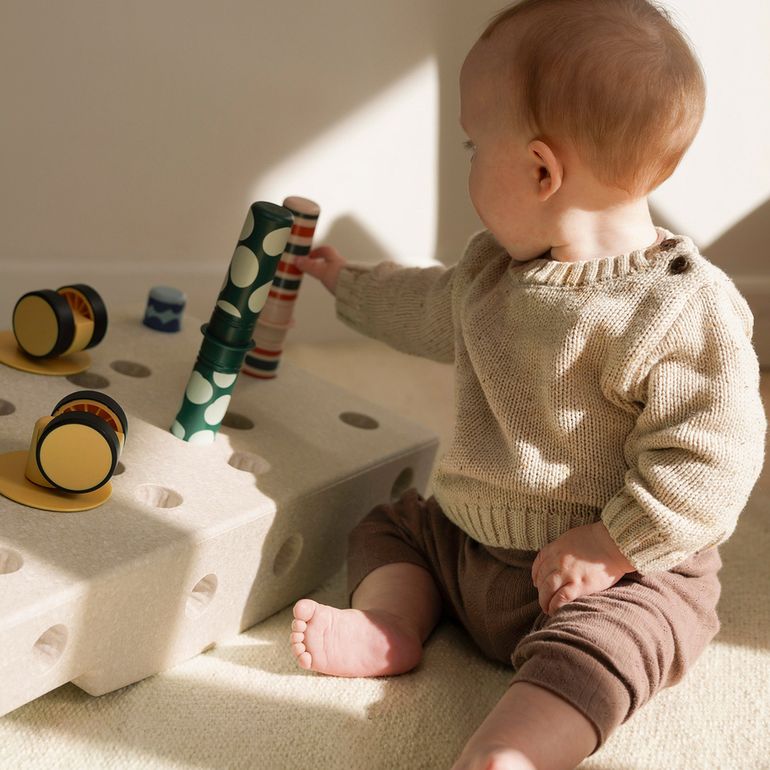 Baby speelt met MODU Activity Set: zittend op beige tapijt grijpt peuter naar groen-wit rammelaar tussen gele en zwarte cilinders in zandgrijs schuims