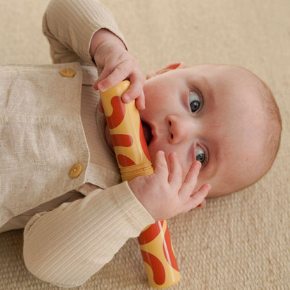 MODU Baby Activity Set: baby speelt met geel-rode rammelaar vanuit bovenaanzicht, zittend op zandkleurig schuimblok