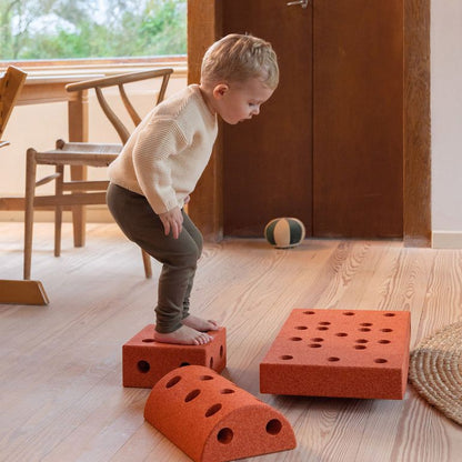 Peuter balanceert op MODU Curiosity Set blokken in woonkamer, speelgoedopstelling met meerdere rode modules op houten vloer