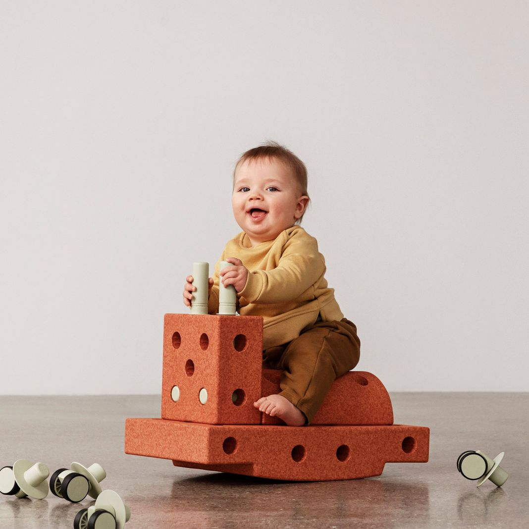 MODU Curiosity Set – peuter zit op gestapelde burnt orange bouwblokken met palen, speelt met houten cilinders, wieltjes op voorgrond