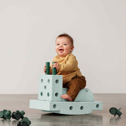 Baby zittend op MODU Curiosity Set bouwblokken in Ocean Mint en Forest Green, met groene wieltjes op de grond