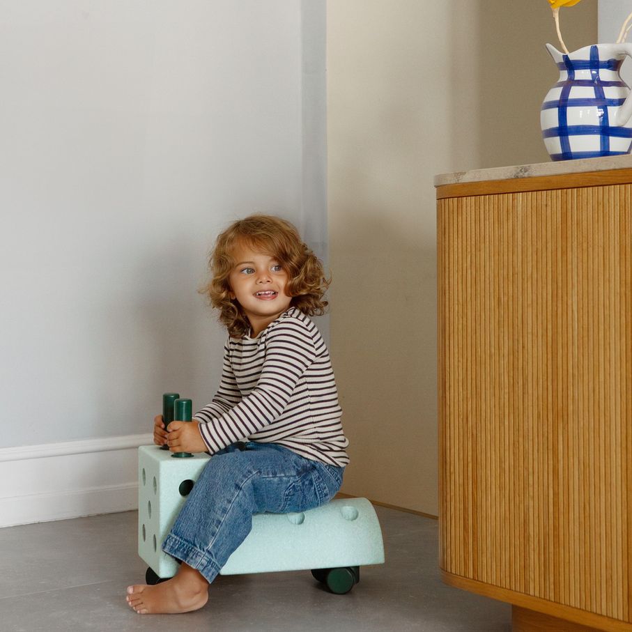 Peuter zit op MODU Curiosity Set Ocean Mint rijdend speelgoed in neutrale kamer met houten kast en gele bloem