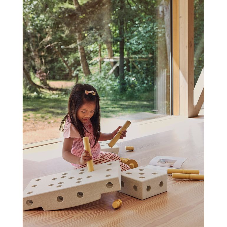 Meisje speelt met MODU Curiosity Set: houten bouwblokken met gaten en houten wielen op houten tafel, uitzicht op groene tuin