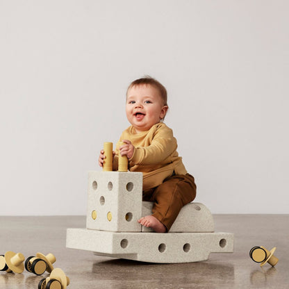 MODU Curiosity Set Sand Grey Honey Yellow: peuter zit spelend op gestapelde bouwblokken met houten peilers, houten wieltjes op voorgrond