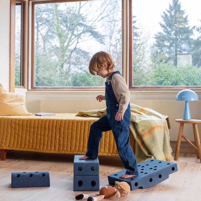 Kind springt op MODU Dreamer Set blokken in woonkamer met gele bank en grote ramen, beweegspeelgoed opstelling