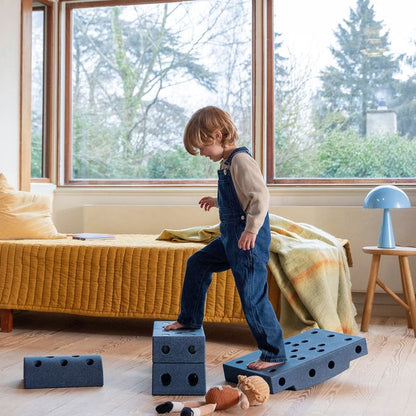 Kind springt op MODU Dreamer Set blokken in woonkamer met gele bank en grote ramen, beweegspeelgoed opstelling