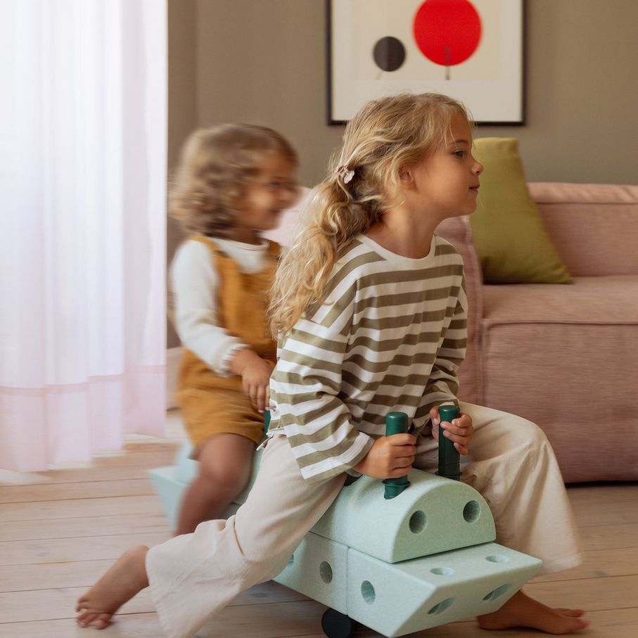 MODU Dreamer Set Ocean Mint: meisje rijdt op modulair bouwsysteem met schuimblokken en wielen, tweede kind speelt op achtergrond