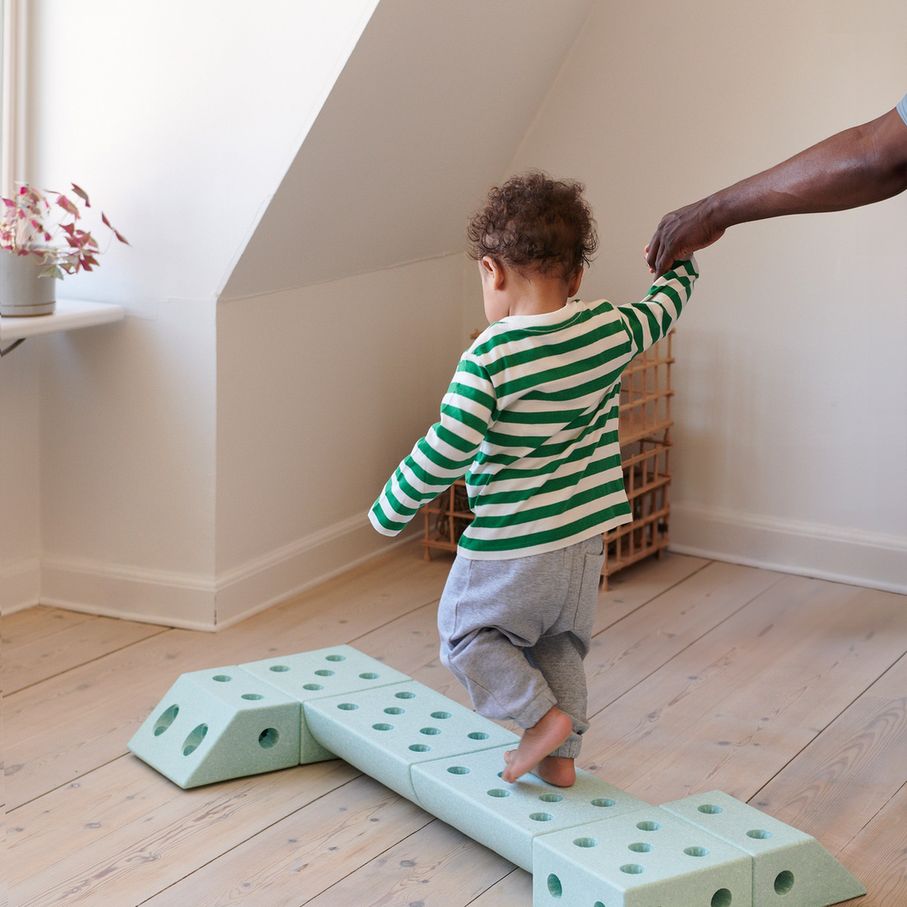 Klein kind in groen-wit gestreept shirt speelt met MODU Dreamer Set modulaire schuimblokken op houten vloer, volwassene helpt bij het spel