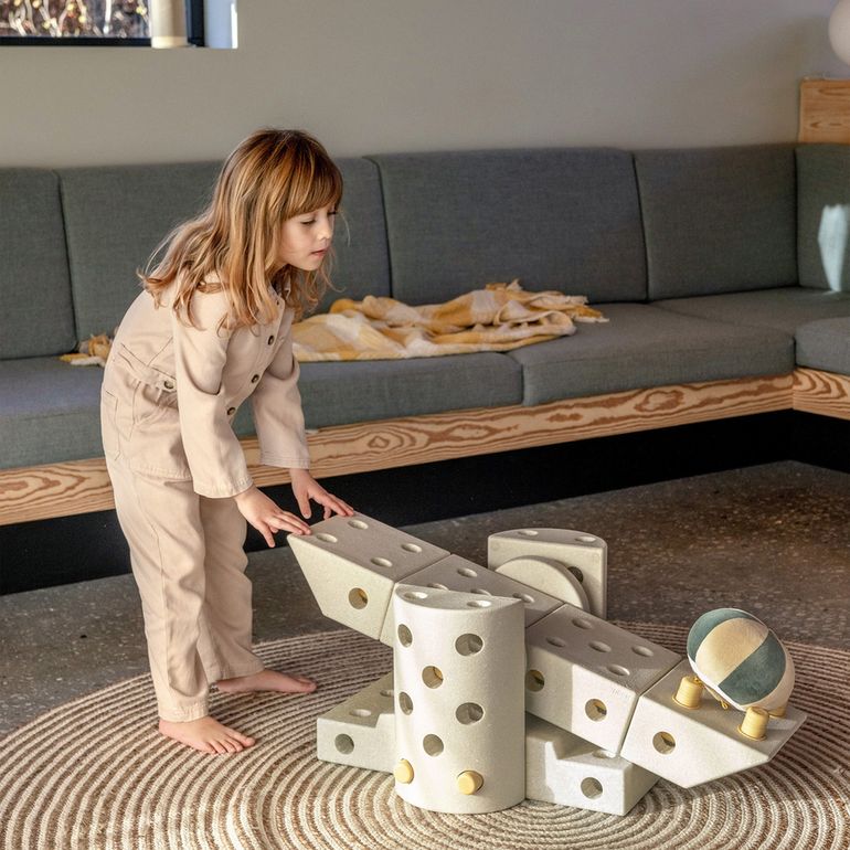 Kind speelt met MODU Dreamer Set bouwblokken op gestreept tapijt, creëert speeltoestel met sand grey en honey yellow onderdelen in moderne woonkamer