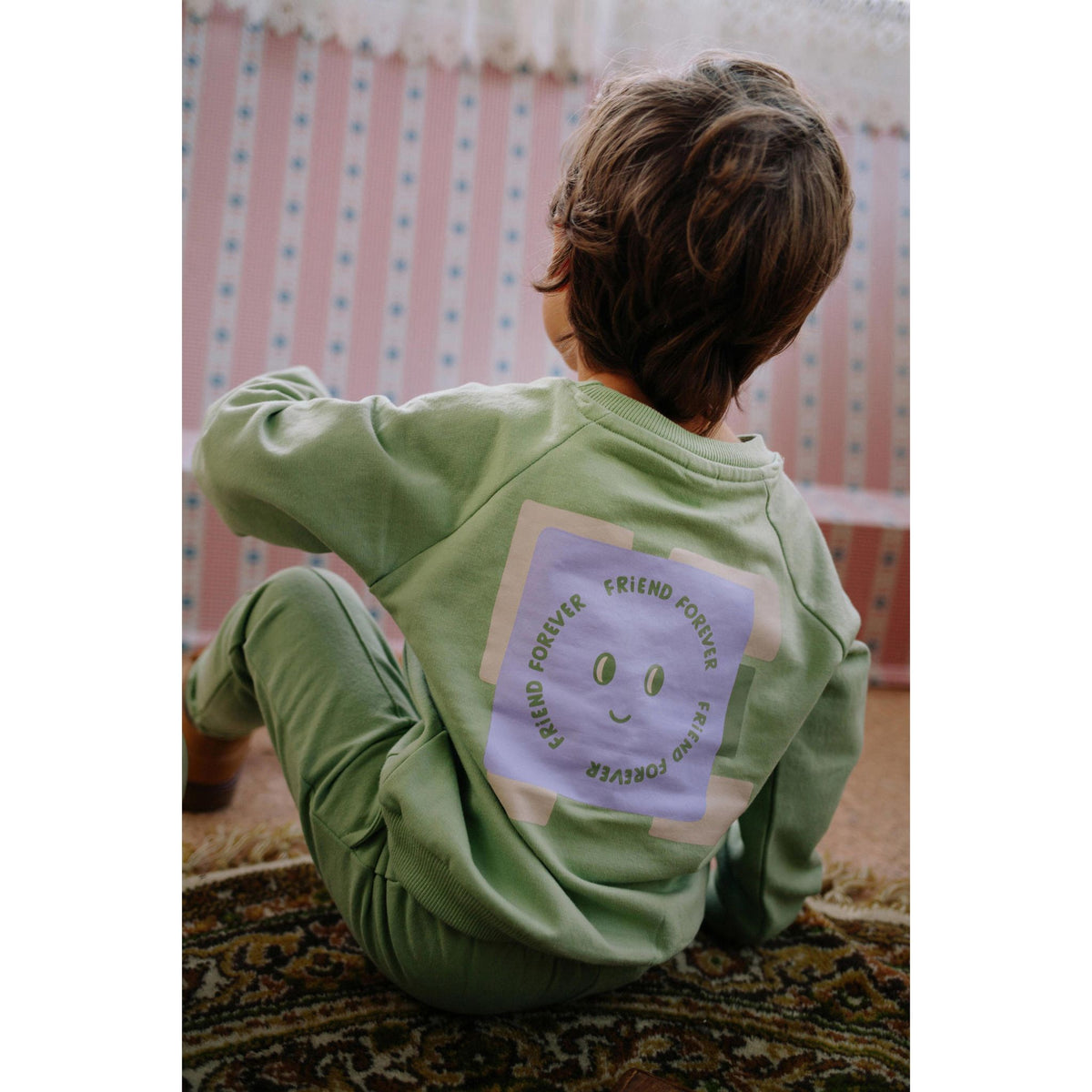 Kind van achteren in groene Riffle Amsterdam hoodie met paars 'Forever Friend' print op de rug, zittend op tapijt