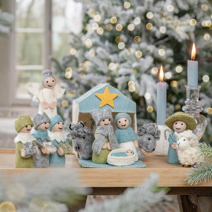 Sjaal met Verhaal kerststal wolvilt ijsblauw-wit met 11 figuren op houten tafel, vooraanzicht met brandende kaars en kerstboom op achtergrond
