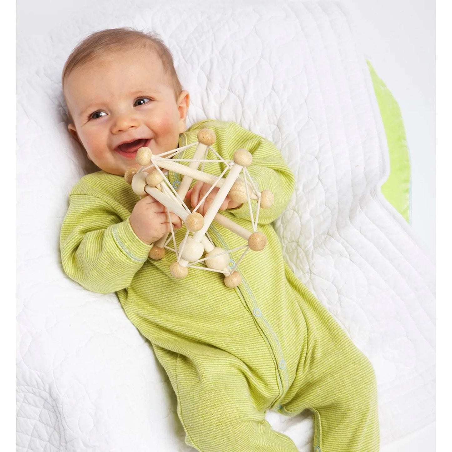 Manhattan Toy Skwish Natural rammelaar in handen van lachende baby in geel pak, bovenaanzicht
