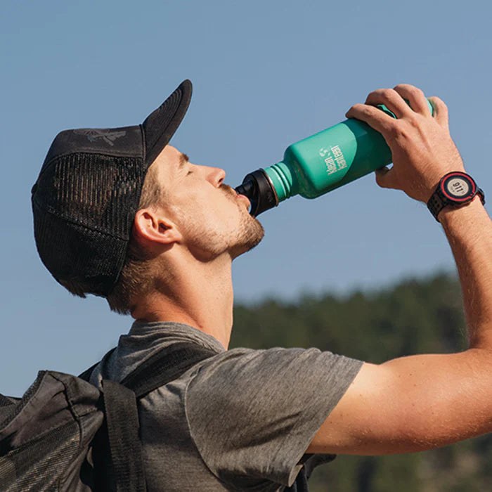 Man drinkt uit groene Klean Kanteen sport cap tijdens outdoor activiteit, zijaanzicht tegen blauwe hemel